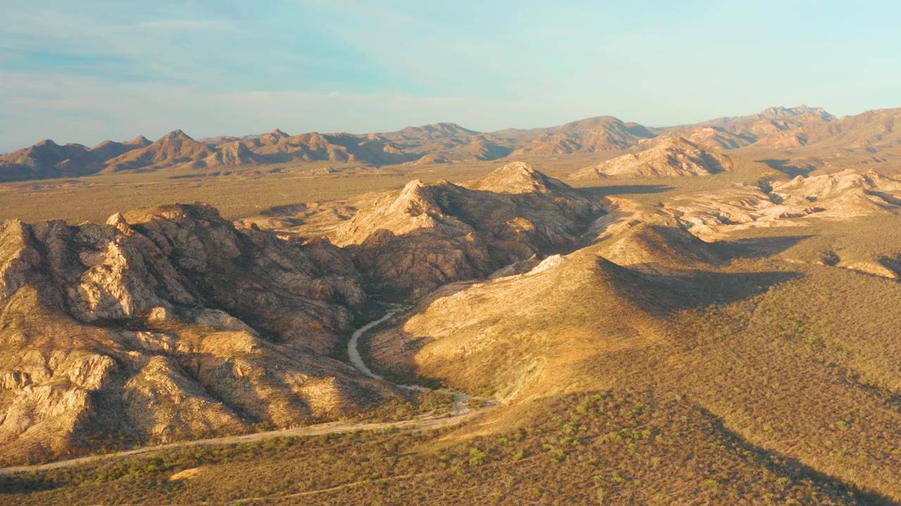 Wide aerial view of Cabo Pulmo's arid mountain landscape, with the road crossing the lower hills, layered tan ridges beyond, clear sky, and no visible people.