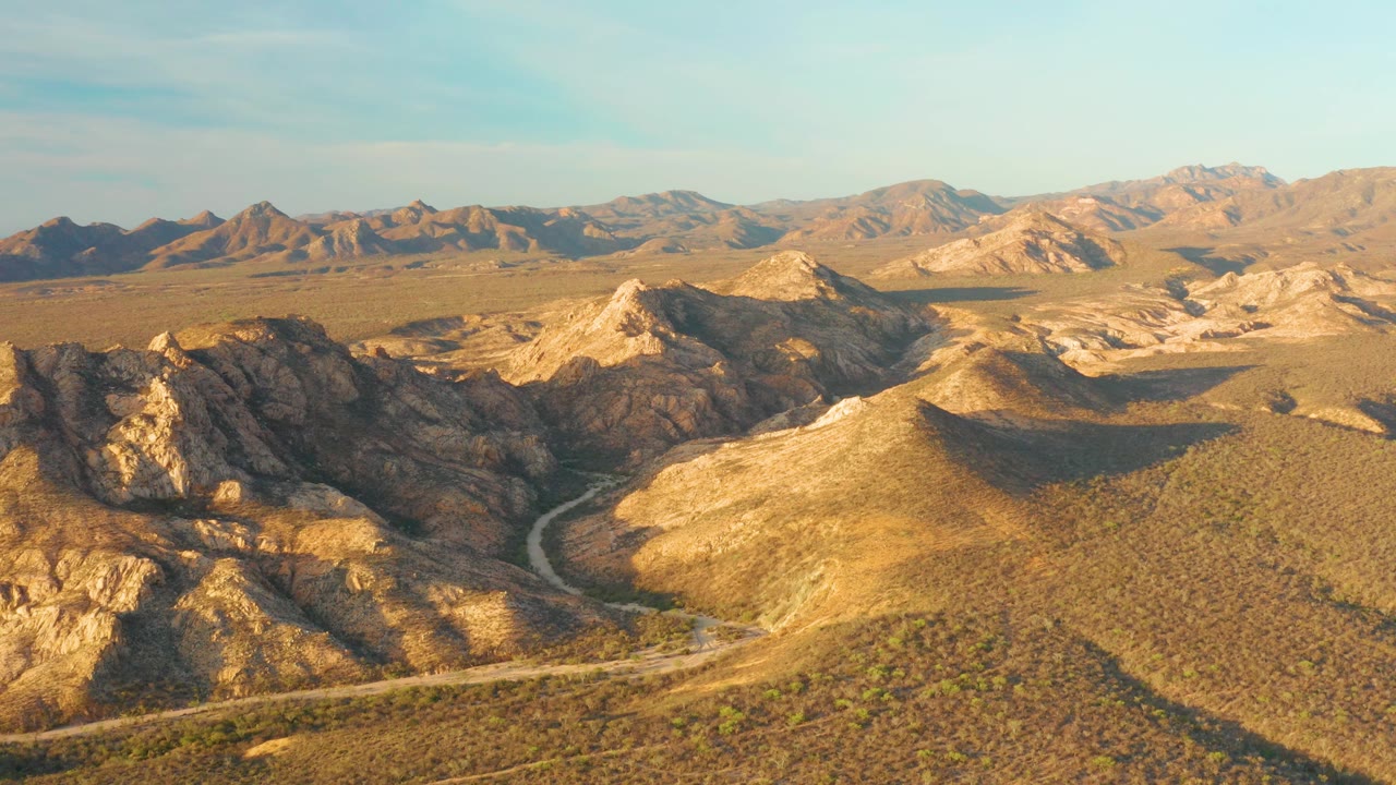 Aerial view over Cabo Pulmo desert terrain, with the winding road tracing the lower frame, rugged foreground rock, sunlit tan hills, distant mountains, and no visible people.