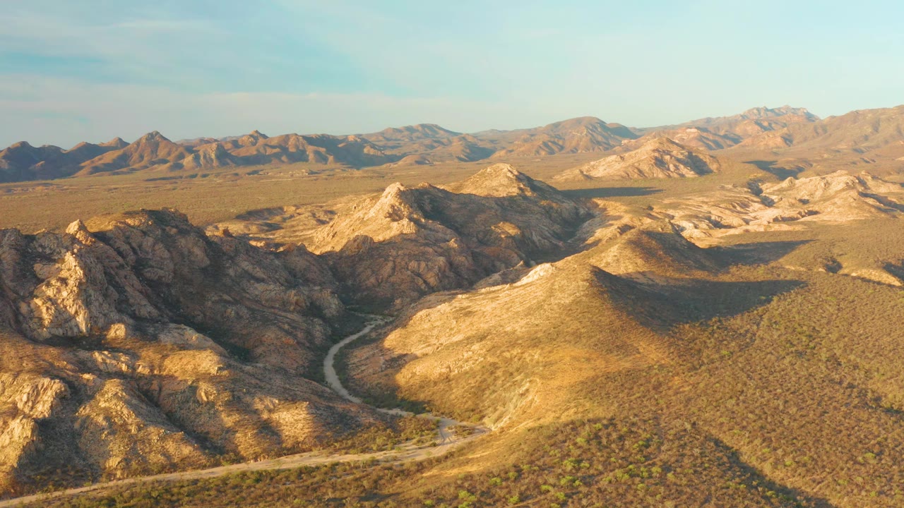 Wide aerial frame over Cabo Pulmo desert terrain, showing the road bending through tan brush, dark rocky ridges, layered mountains, and no visible people.