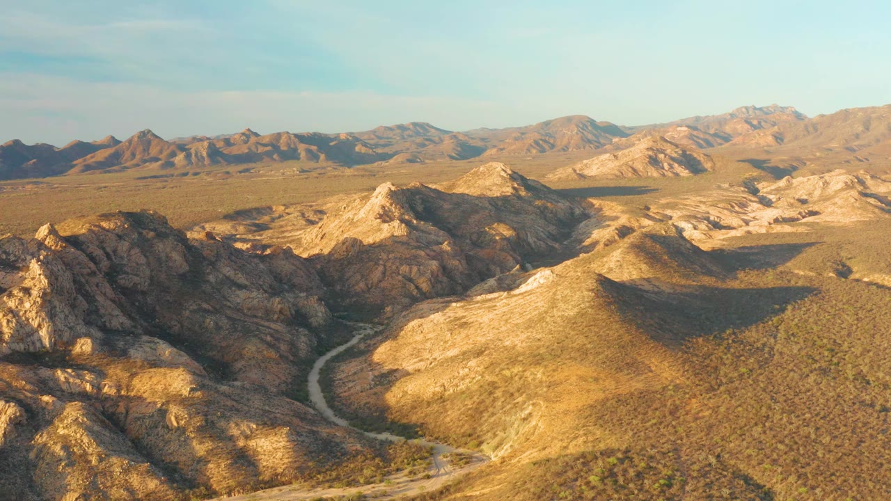 Elevated view of Cabo Pulmo arid hills, where the road curves through scrub-covered terrain below sunlit ridges and distant mountain ranges, with no visible people.