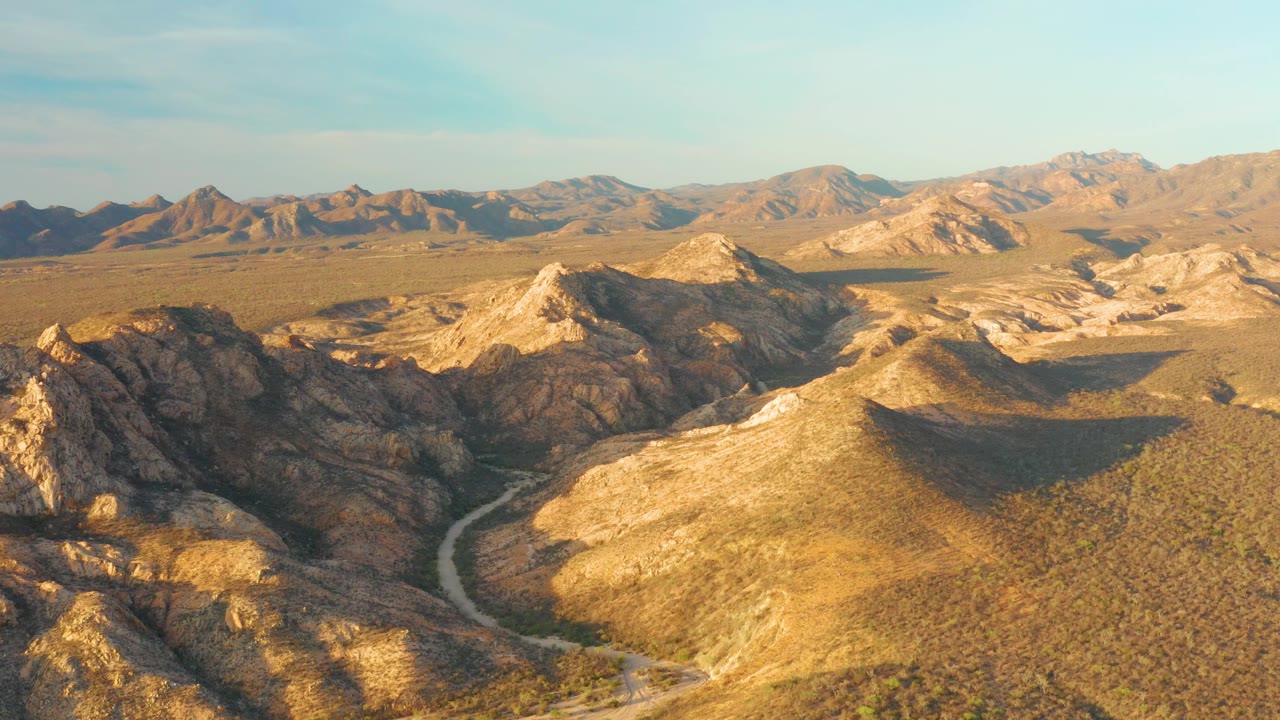 Aerial view of Cabo Pulmo dry mountain terrain, with the road bending through the lower left valley and broad ridges stretching toward the horizon, no visible people.