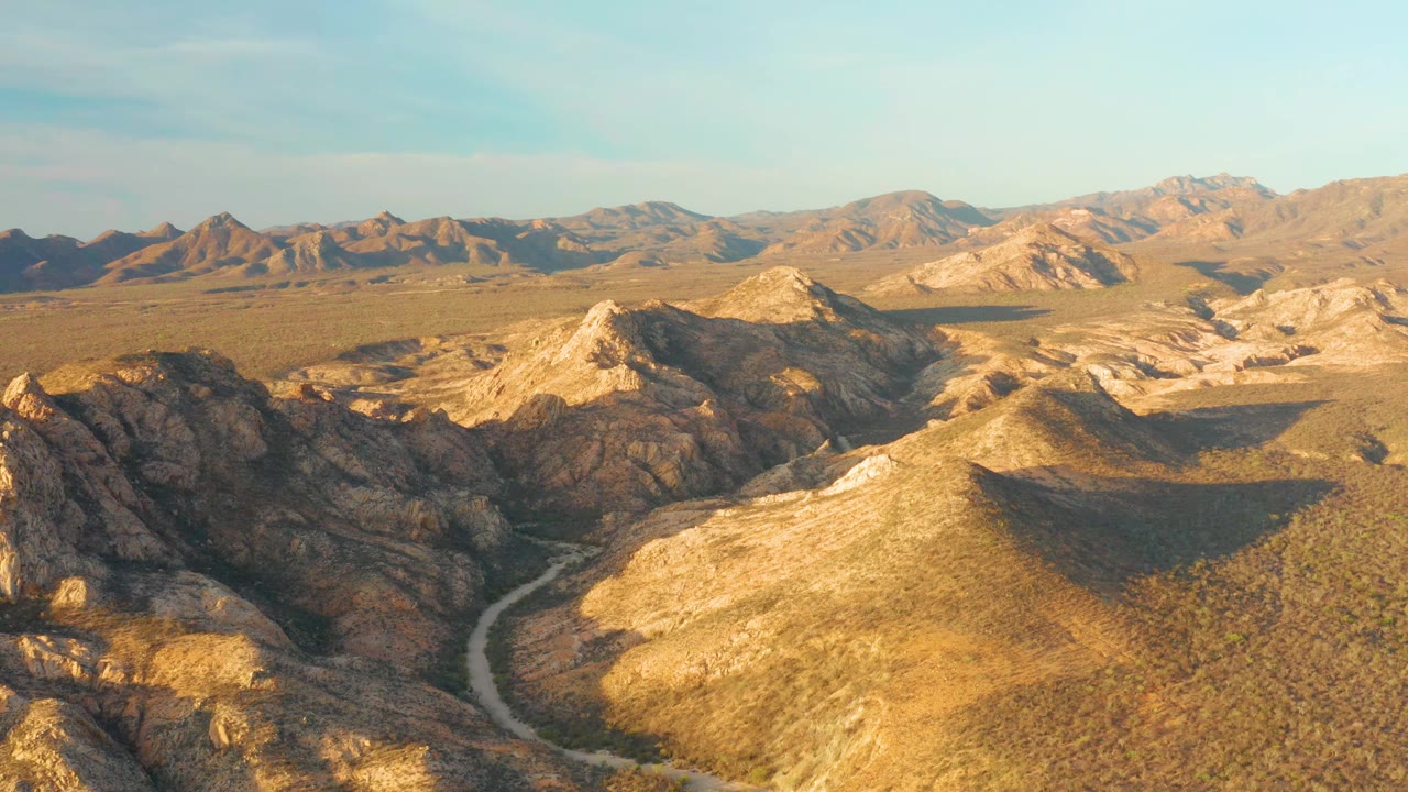 Aerial frame in Cabo Pulmo, Baja California Sur, with the road bending between dark rocky ridges and pale sunlit slopes, no visible people.