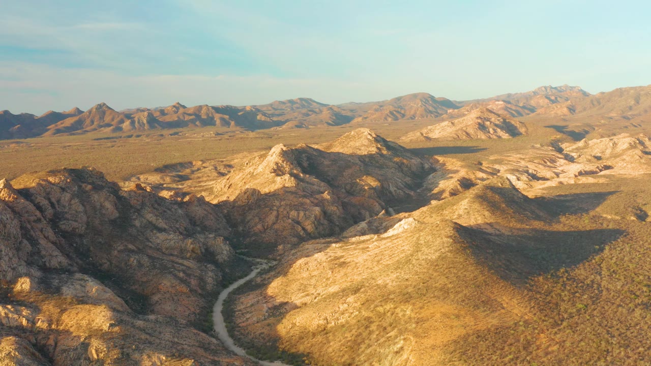 Mid-clip aerial over Cabo Pulmo, Baja California Sur, with a pale road threading past shadowed rock formations and dry golden hills, no visible people.