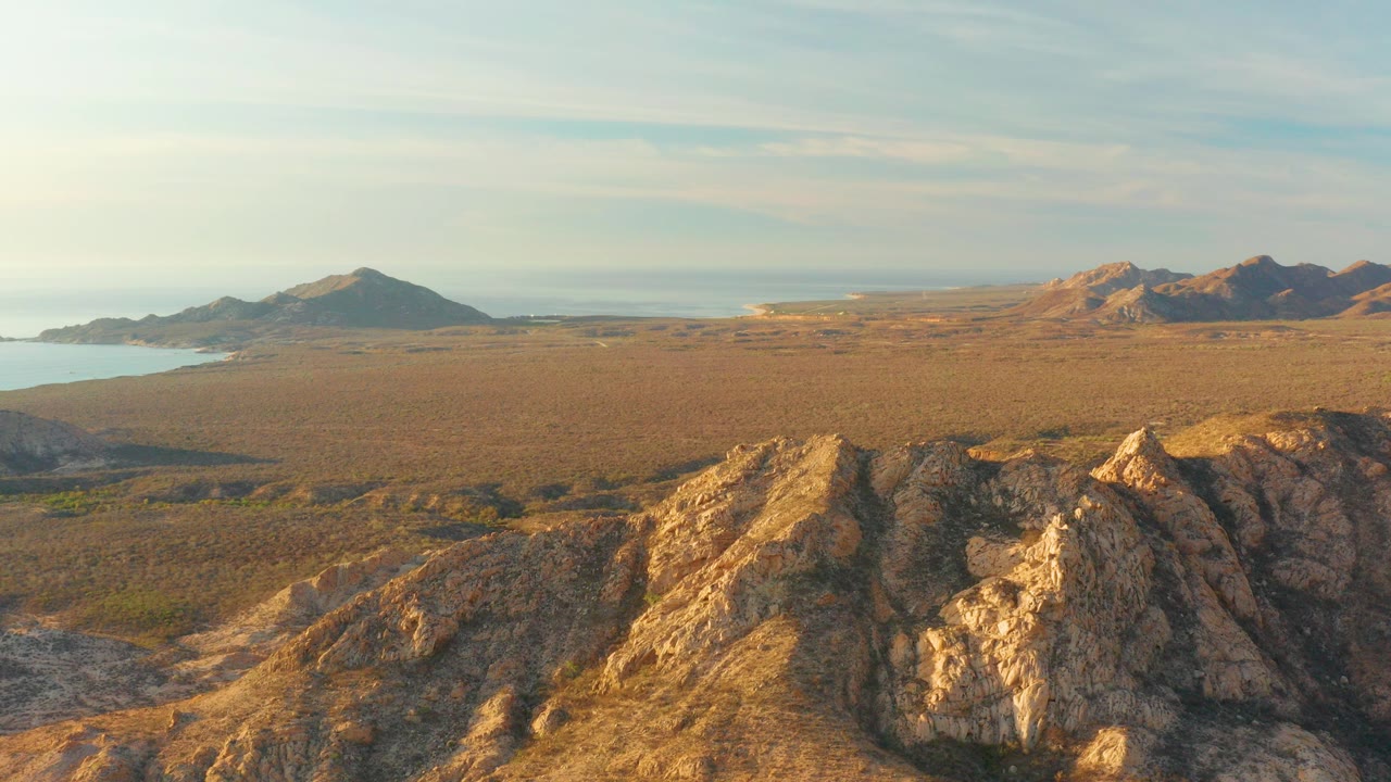 Opening aerial frame over Cabo Pulmo rocky desert ridges, with a wide dry coastal plain, distant hills, a small sea edge at left, pale clouds, and no visible people.