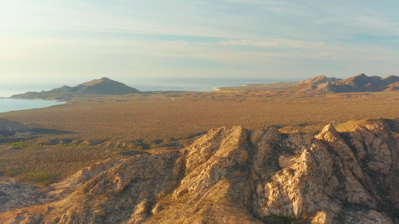 Cabo Pulmo aerial frame with sunlit angular rocks in front, flat coastal desert beyond, darker hills at left, brighter rocky hills at right, distant sea on the horizon, and no visible people.