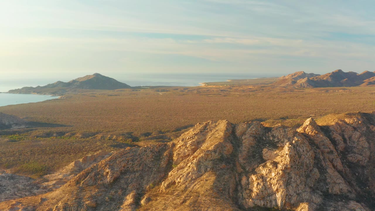 Wide aerial composition of rocky ridge, dry plain, distant hills, and sea edge.