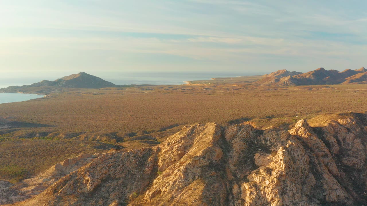 Cabo Pulmo aerial view over golden rocky ridges, wide arid plain, distant mountains, ocean at the left edge, and no visible people.