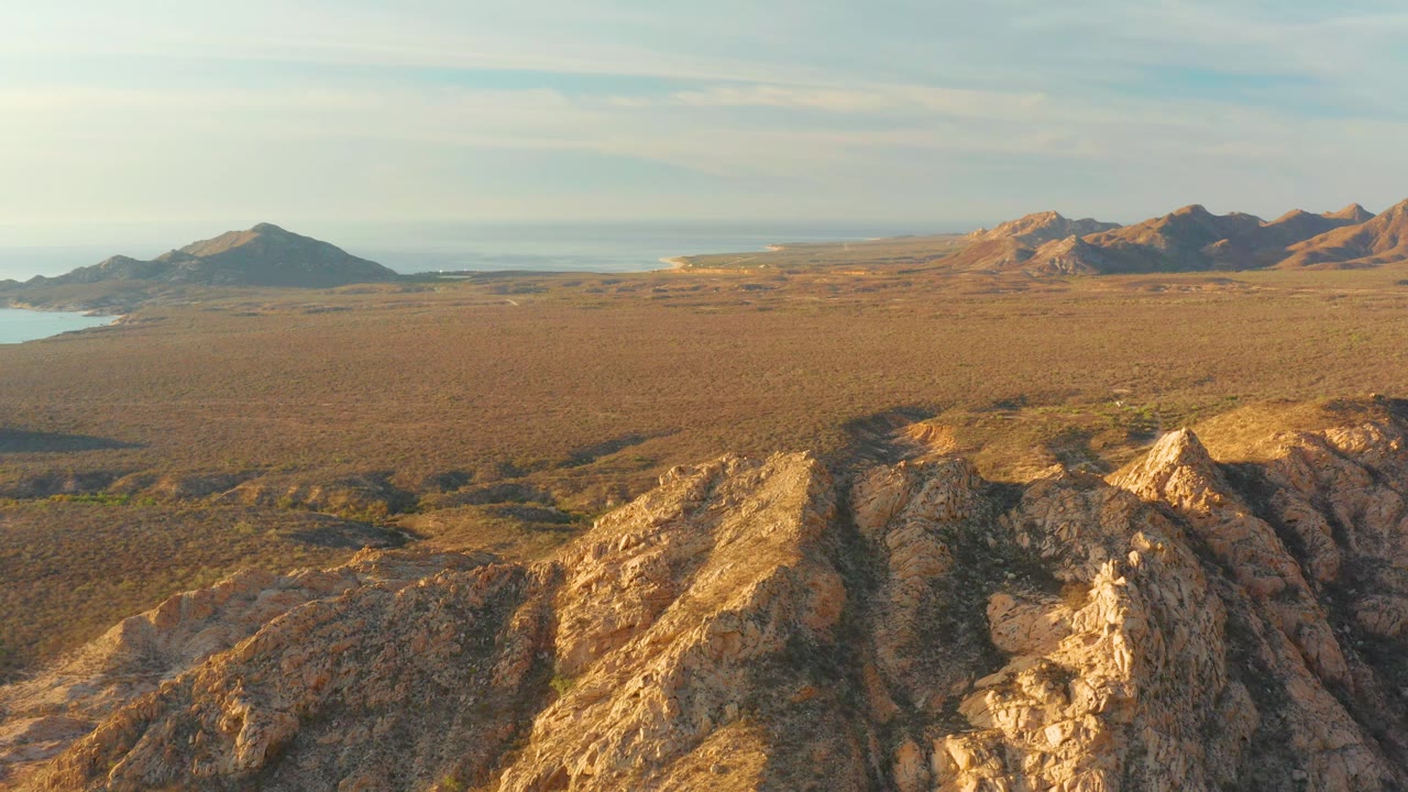 Aerial frame over Cabo Pulmo showing jagged golden ridges in the foreground, a flat arid valley across the center, mountain ranges in the distance, pale water near the left edge, and no visible people.