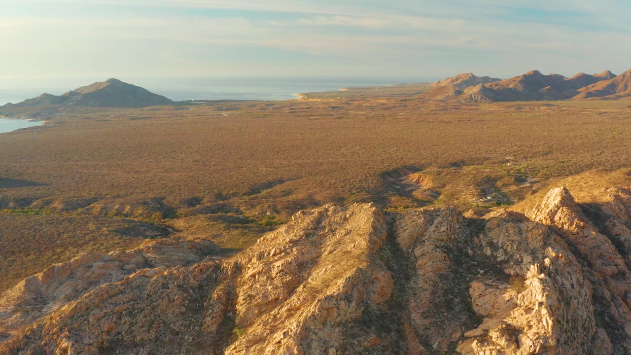Aerial view in Cabo Pulmo, Baja California Sur, with sunlit rocks in the foreground, a wide dry plain, distant mountains, a small band of water at left, and no visible people.
