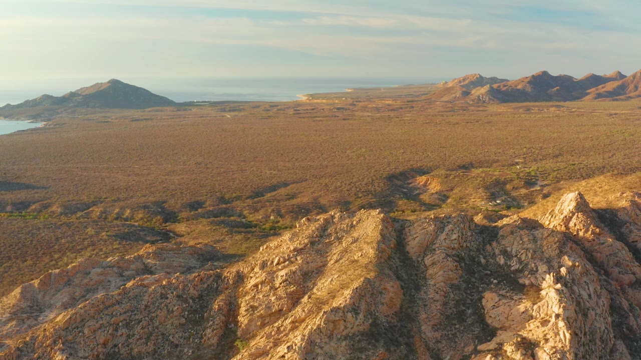 Aerial view over Cabo Pulmo terrain with golden rock formations in front, scrub-covered flatland beyond, distant mountains to the right and left, and no visible people.