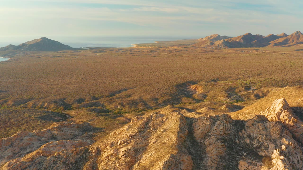 Wide aerial view in Cabo Pulmo, Baja California Sur, with sunlit rocky foreground hills, a dry open plain, distant mountains, light clouds, and no visible people.