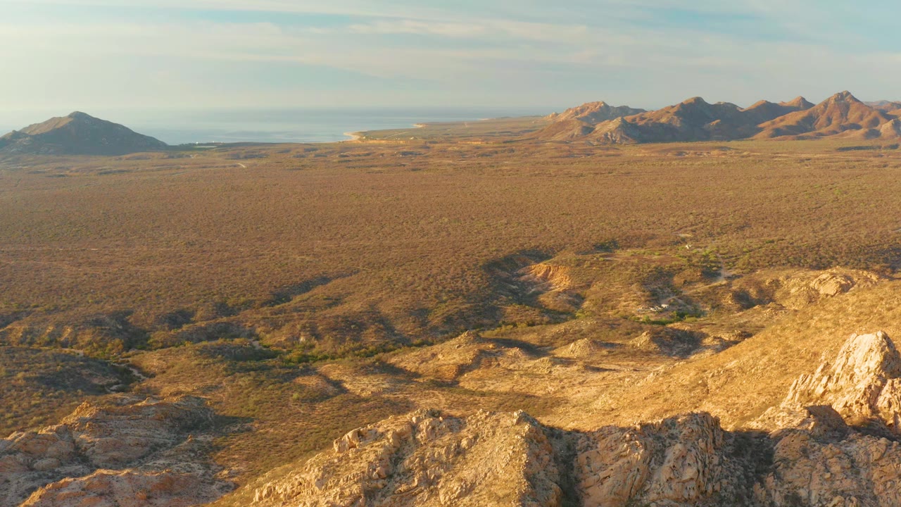 Early frame in Cabo Pulmo, Baja California Sur, with bright rock outcrops in the foreground, brown valley scrub, distant hills, and no visible people.