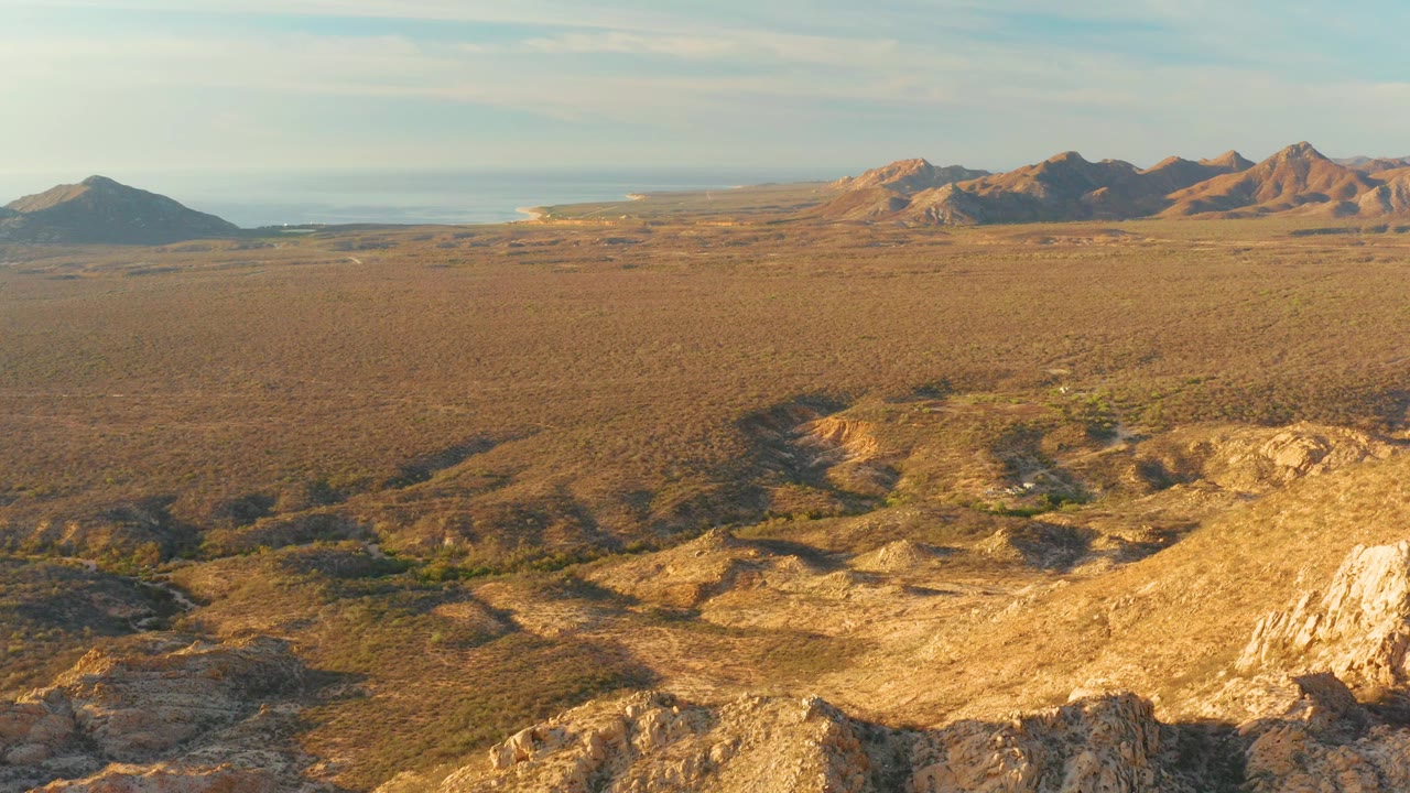 Mid-clip Cabo Pulmo view with shadowed foreground rock, open dry valley, low green patches, distant hills, blue sky, and no visible people.