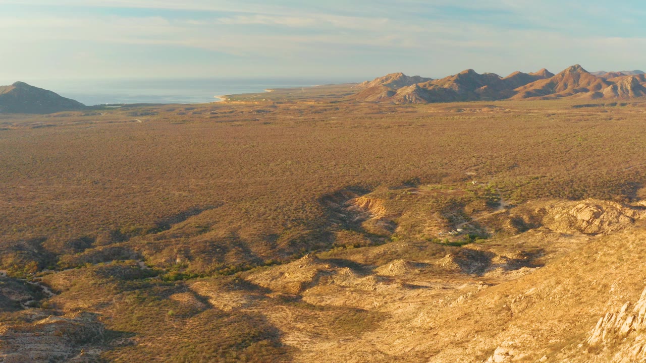 Aerial frame from Cabo Pulmo, Baja California Sur showing tan rocky foreground, wide scrub plain, distant mountains, pale sky, and no visible people.