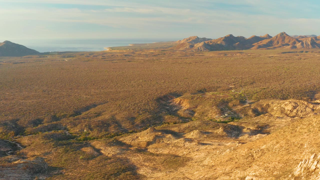 Aerial frame over Cabo Pulmo desert terrain with sunlit foreground rocks at lower right, dense scrub across the plain, distant mountains, and no visible people.