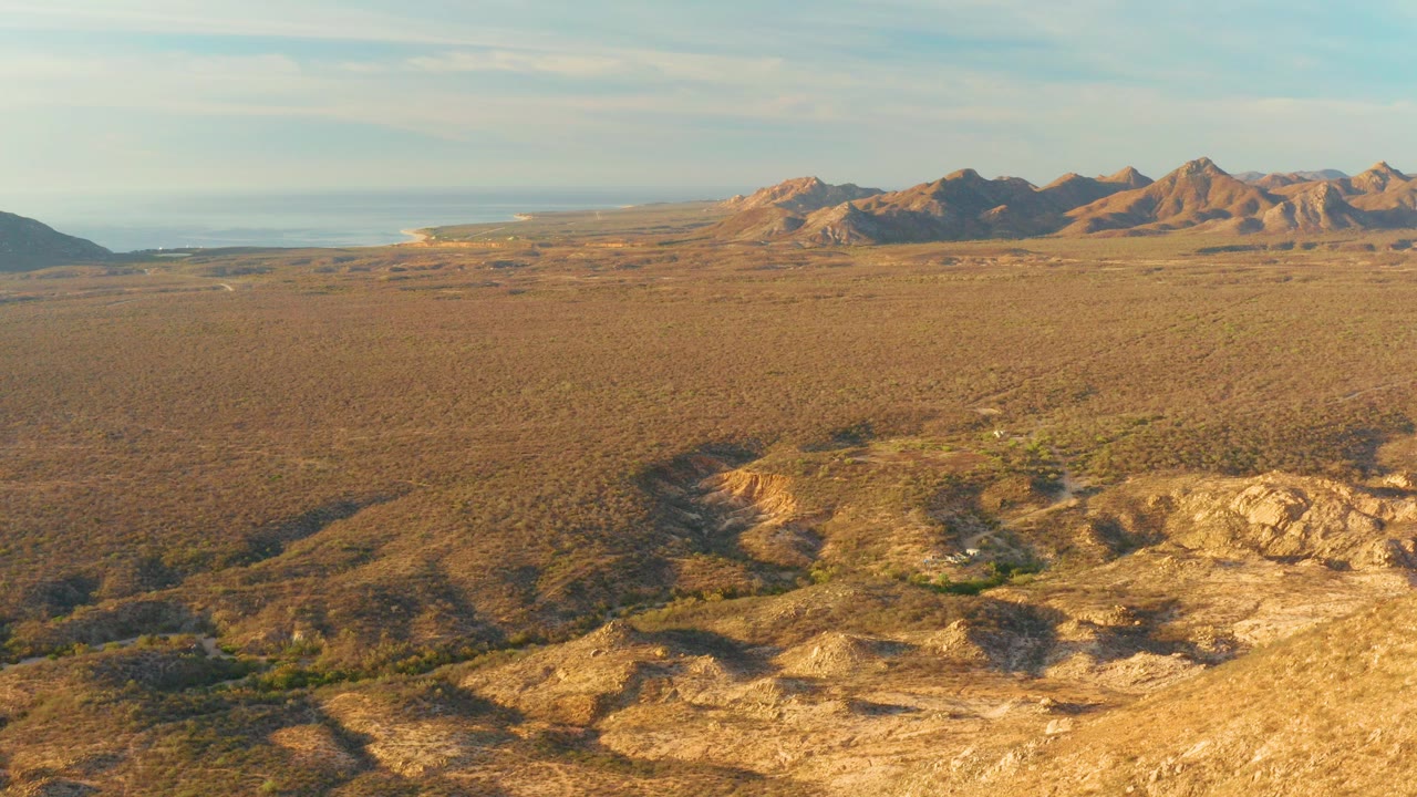 Wide aerial frame over Cabo Pulmo desert terrain with sunlit rocky ridges in front, scrub-covered plain beyond, distant mountains, pale sky, a faint waterline at left, and no visible people.