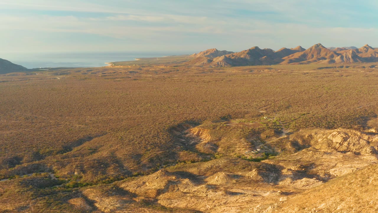 Aerial frame over Cabo Pulmo with uneven rocky desert in the lower foreground, a wide scrub plain through the center, distant mountain ridges, bright sky, and no visible people.