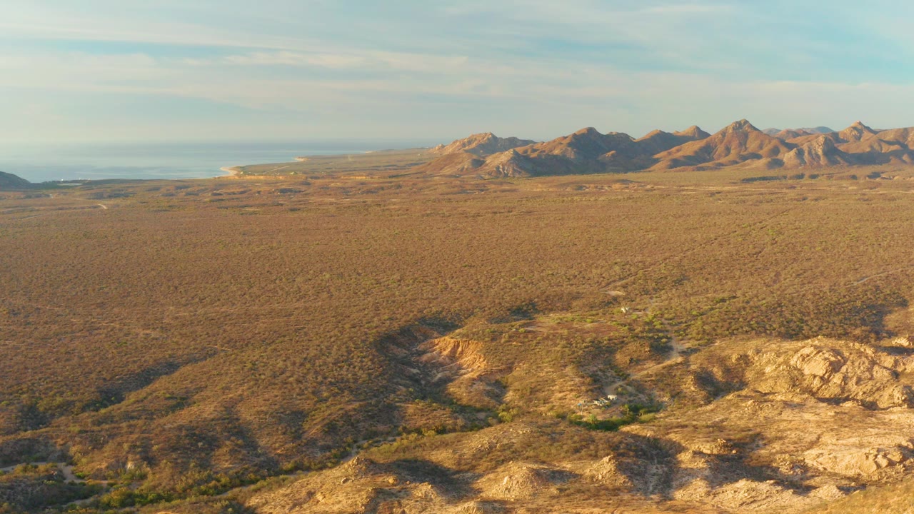 Wide aerial frame over Cabo Pulmo desert plain with rocky foreground ridges, sparse scrub, distant mountain ranges, thin clouds, and no visible people.