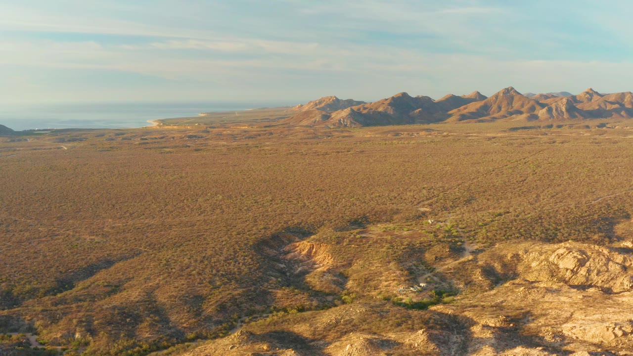 Wide desert aerial with mountains, scrubland, and rocky foreground shadows.