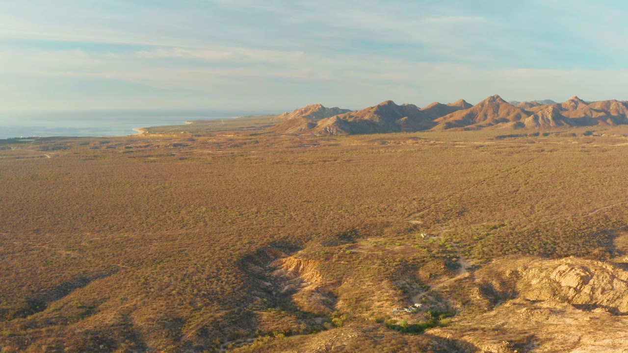 One second in, the Cabo Pulmo aerial view keeps the eroded rocky foreground prominent below a broad brown plain, with distant mountains, pale sky, ocean at left, and no visible people.