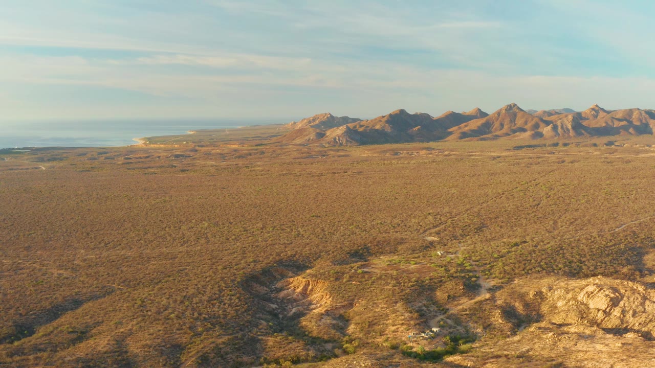 At 0:03, a rocky wash and sunlit tan ground sit below a wide Cabo Pulmo desert plain, with mountains in the background, ocean visible left of the range, and no visible people.