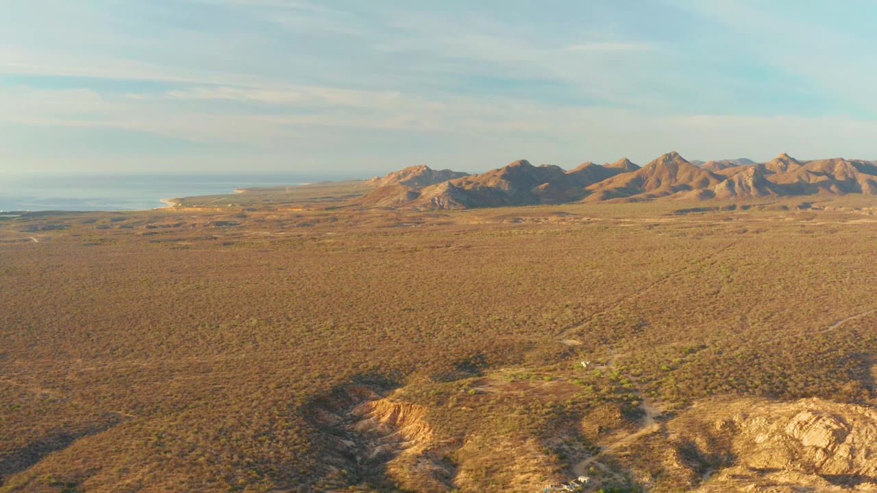 Aerial frame in Cabo Pulmo showing brown scrubland spreading toward sunlit mountains, a thin ocean band at left, and no visible people.