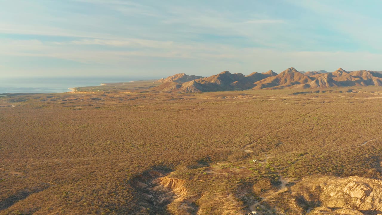 Cabo Pulmo desert terrain fills the frame below a distant ridgeline, with rocky ground in the foreground, ocean on the left horizon, and no visible people.