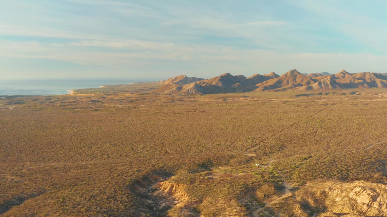 High aerial view over Cabo Pulmo's dry plain, with sparse vegetation below, mountains across the background, ocean faintly at left, and no visible people.