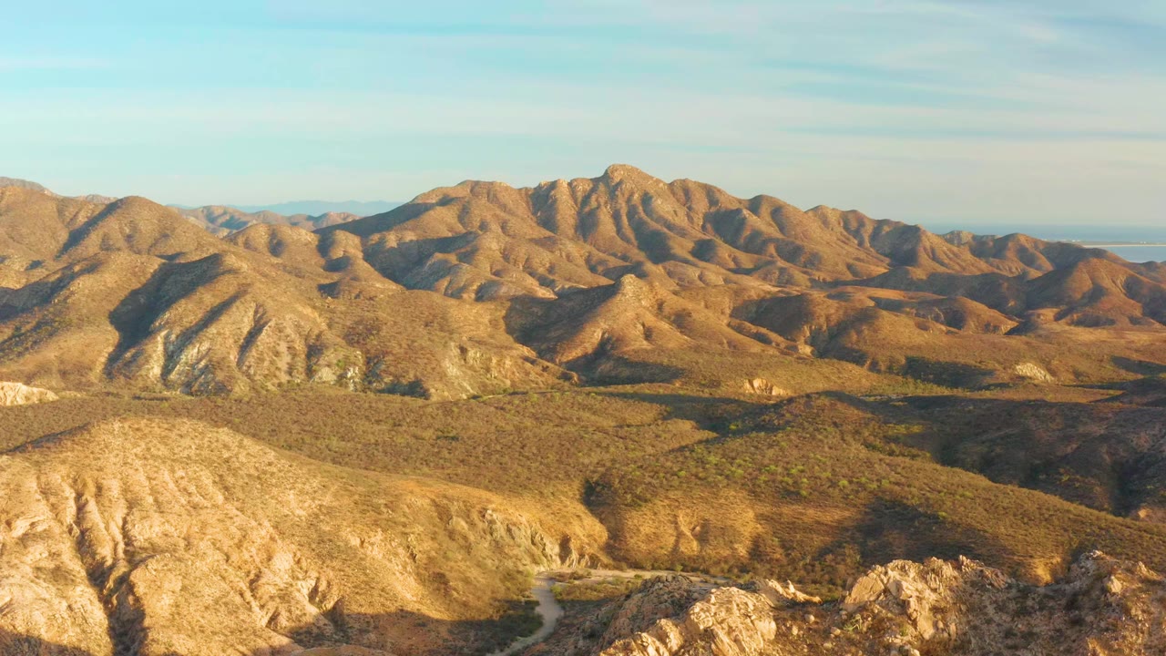 Wide aerial view of dry brown mountain hills near Cabo Pulmo, Baja California Sur, Mexico, with rocky foreground ridges, pale blue sky, and no visible people.