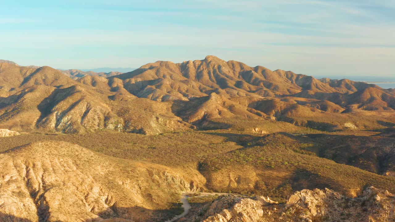 Aerial frame of the Cabo Pulmo mountain range with golden brown slopes, broad terrain shadows, a low distant horizon, and no visible people.