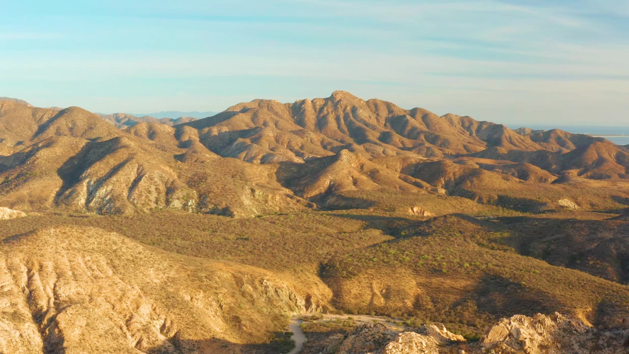 One second in, the Cabo Pulmo landscape holds a broad view of sunlit brown hills, darker scrub patches, the road near the lower center, and no visible people.