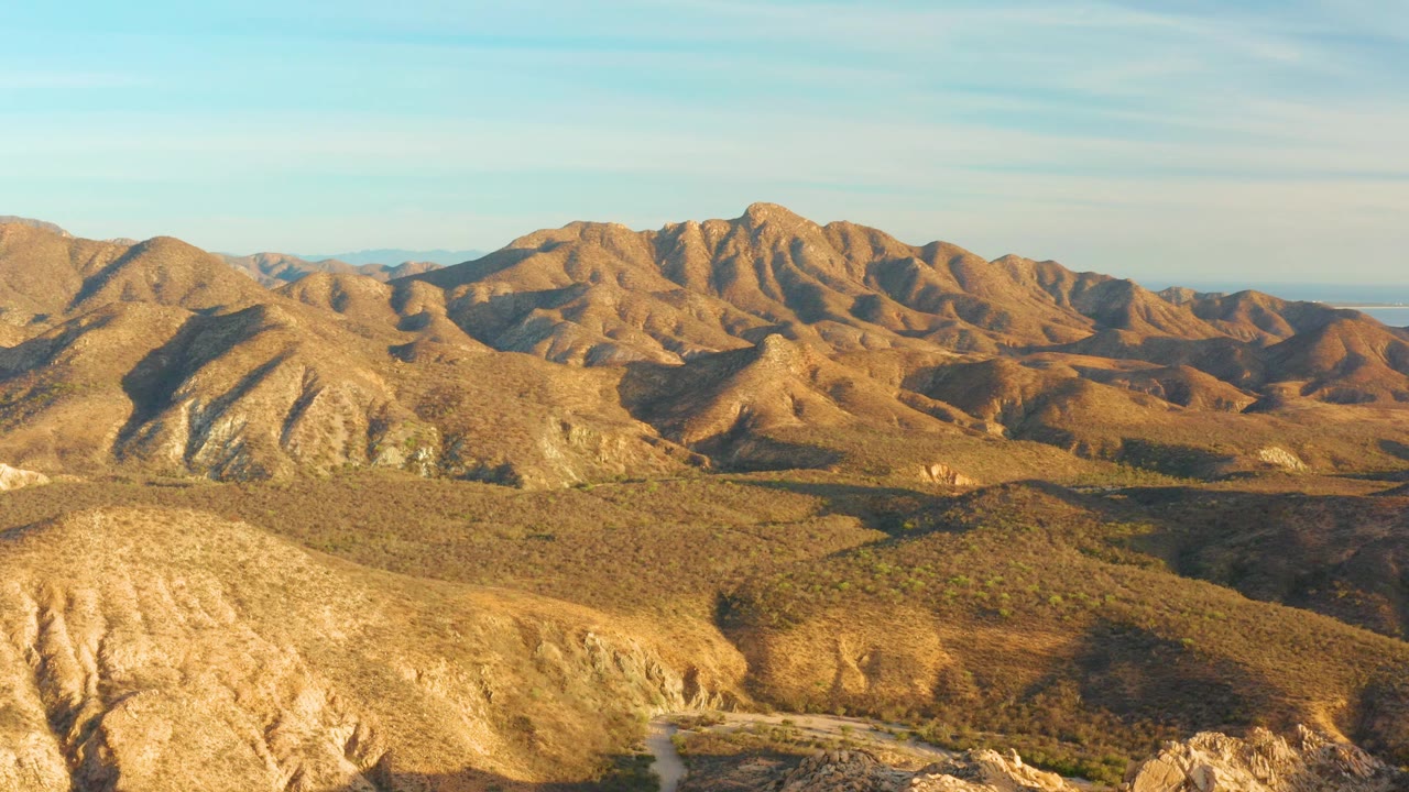 Mid-clip, the aerial view shows a bright foreground ridge, shadowed scrub valley, tan mountain folds beyond, the winding road low in frame, and no visible people.