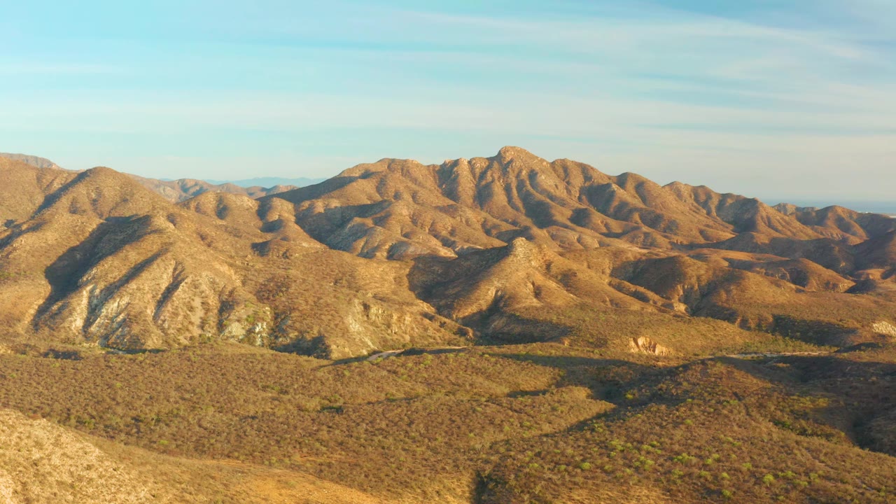Second frame of the Cabo Pulmo ridge line showing sunlit brown slopes, darker shadow bands across the valley floor, pale blue sky, and no visible people.