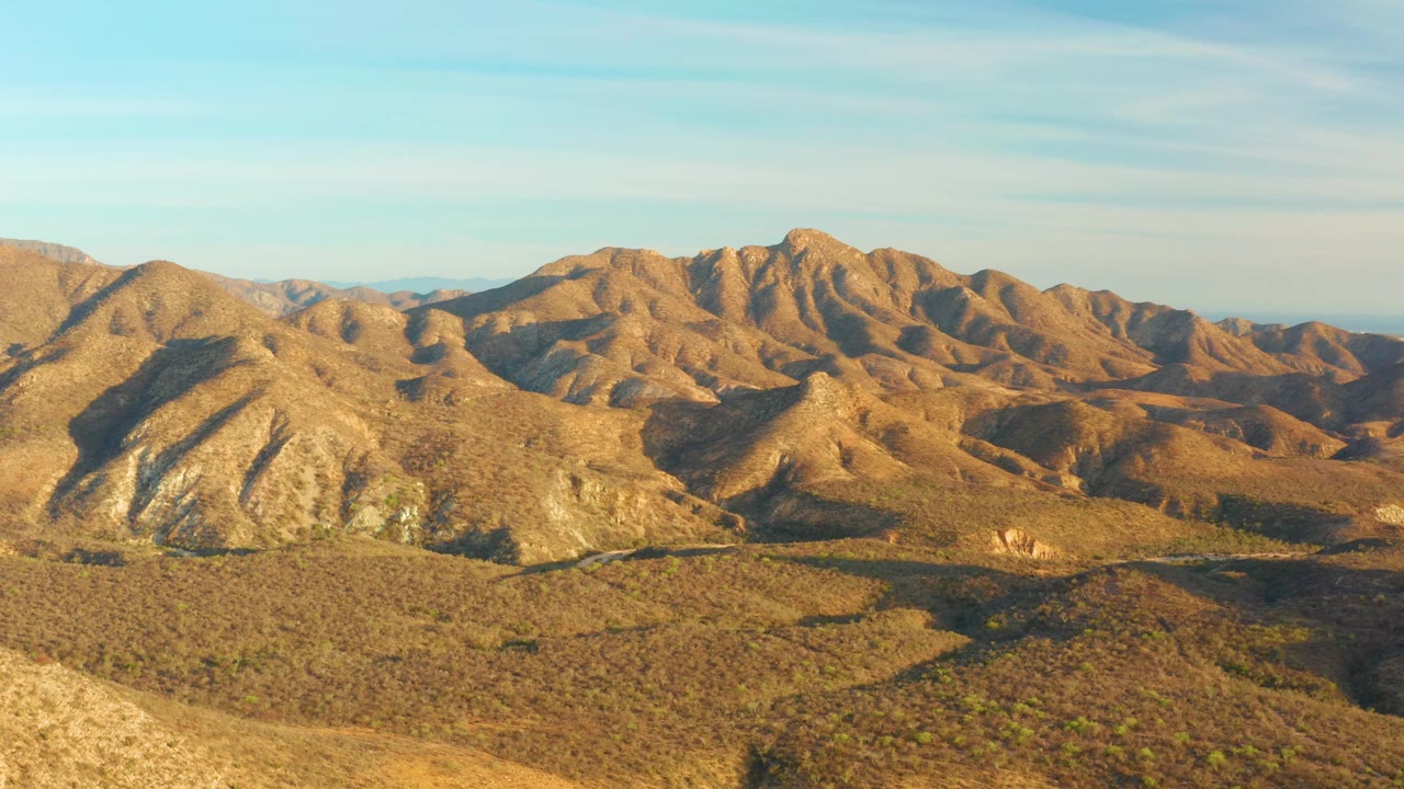 Third frame holds on the Cabo Pulmo desert mountains, with rounded brown ridges across the center, scrubby terrain in front, thin clouds in blue sky, and no visible people.