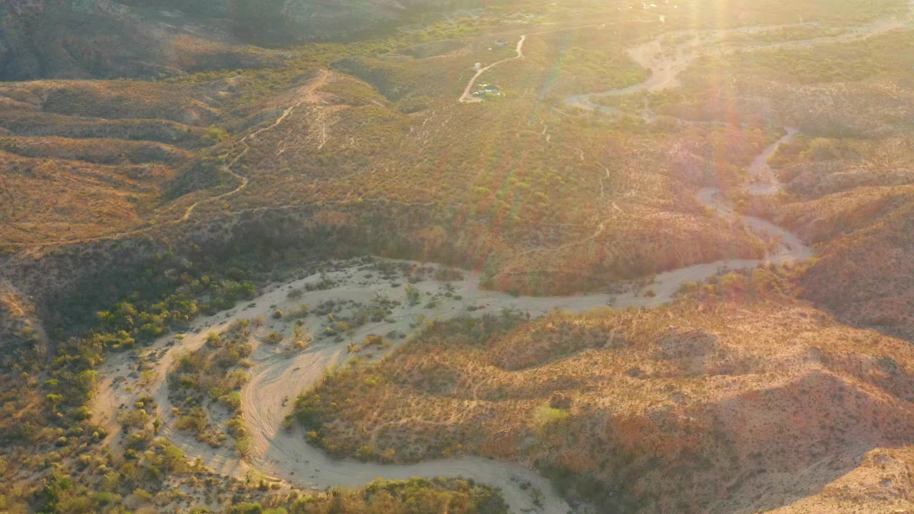 Aerial view of arid hills with a pale winding wash and sun flare.