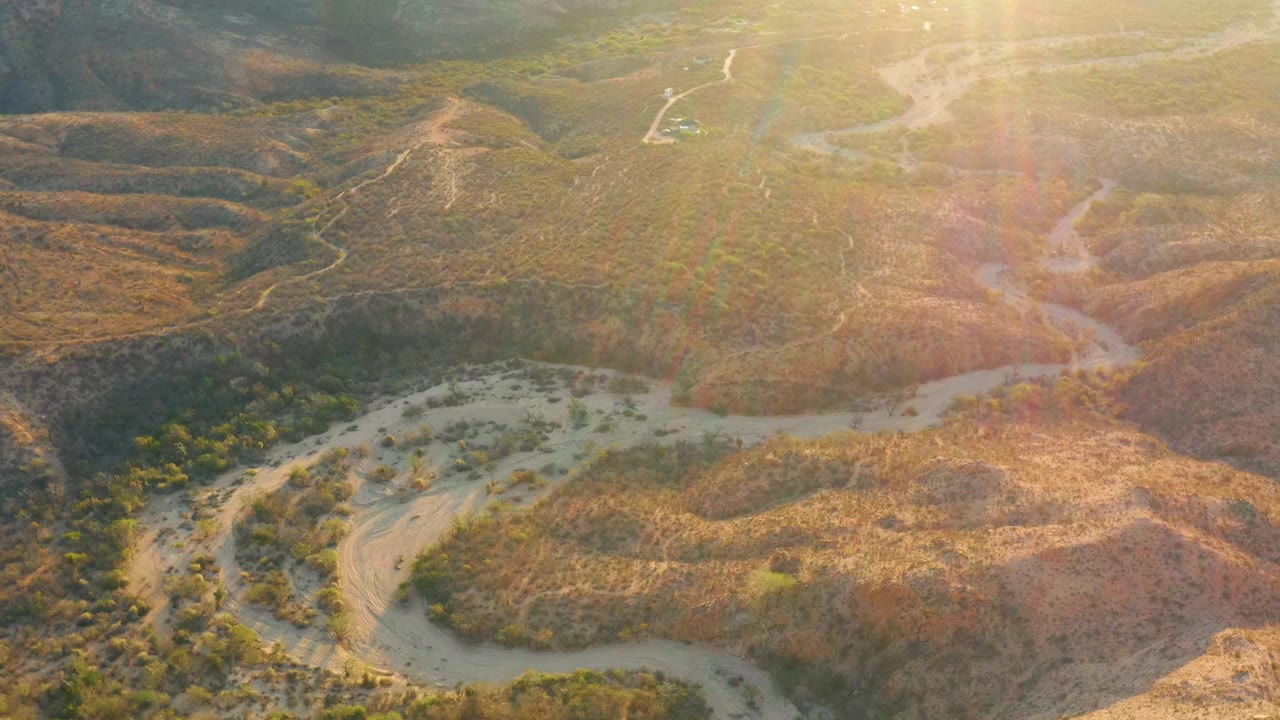 Aerial view in Cabo Pulmo, Baja California Sur with scrub-covered hills, pale drainage curves crossing the lower frame, bright lens flare over the upper right, and no visible people.