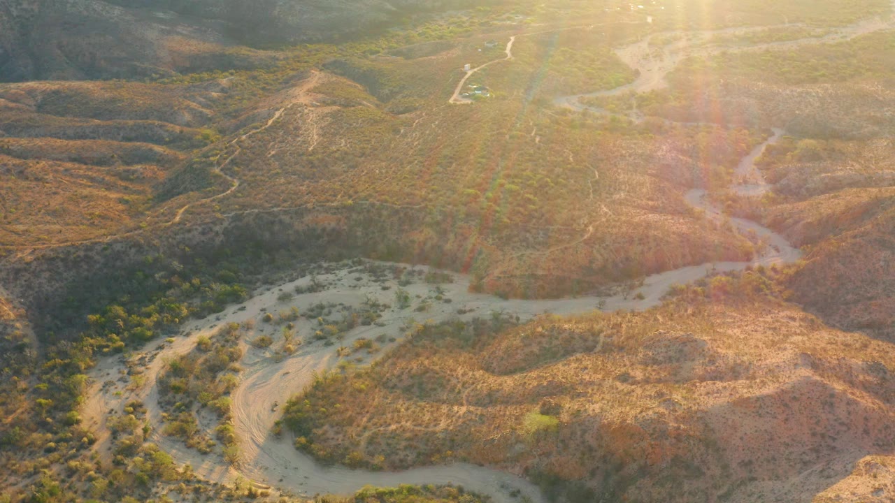 Cabo Pulmo aerial frame showing brown ridges, pale branching dry channels, sparse green vegetation, warm golden glare across the right side, and no visible people.