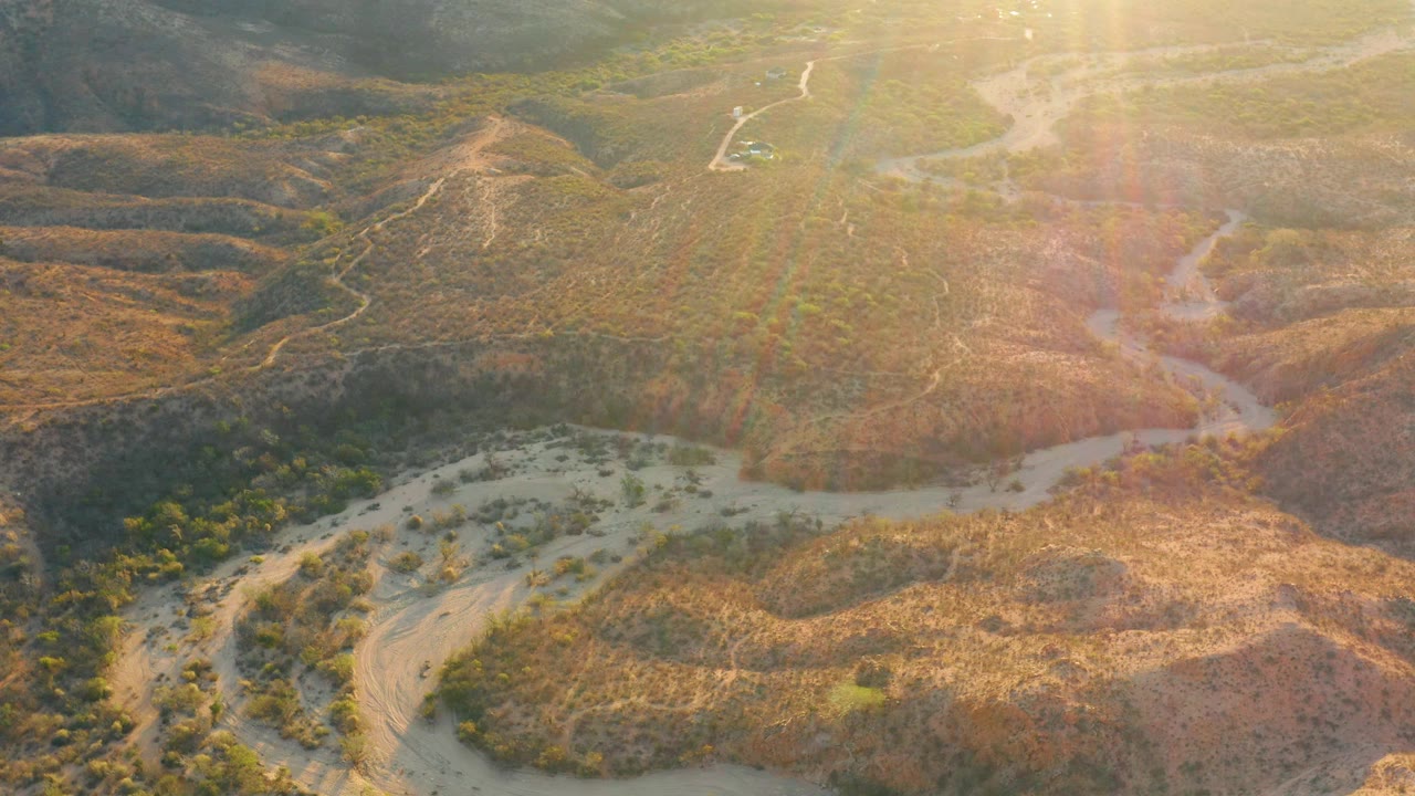 Aerial view over Cabo Pulmo, Baja California Sur with a winding pale wash along the lower left, rounded dry hills through the center, sun streaks over the right half, and no visible people.