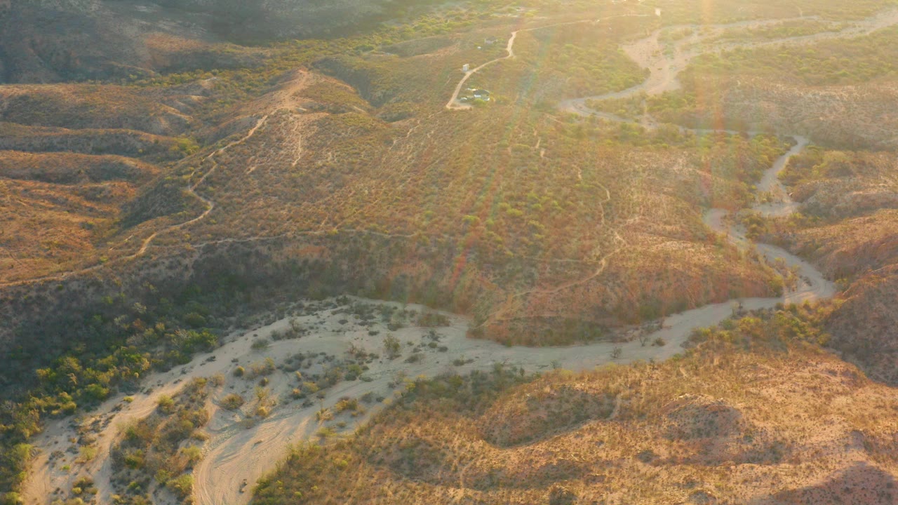 Aerial view of arid hills and a pale winding wash in golden light.