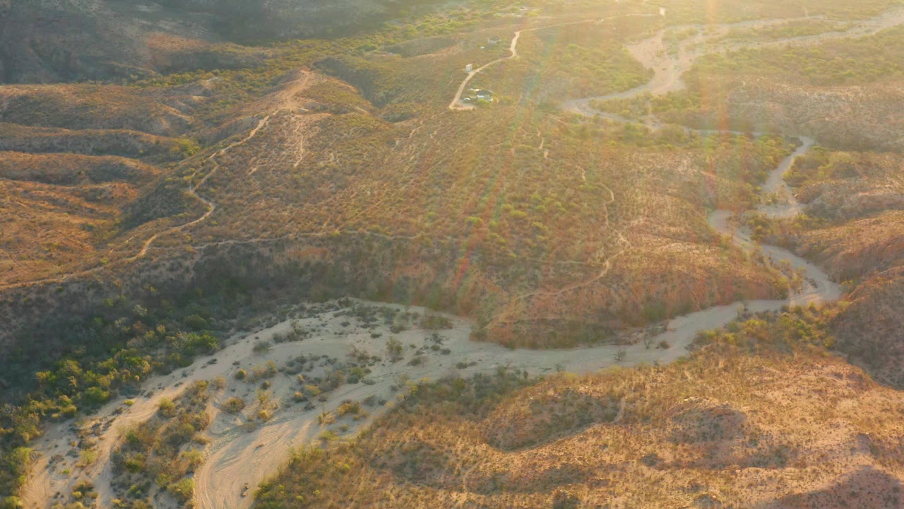 High aerial frame in Cabo Pulmo showing scrub-covered hills, a pale branching wash, faint dirt trails, and sunlit brown terrain, with no visible people.