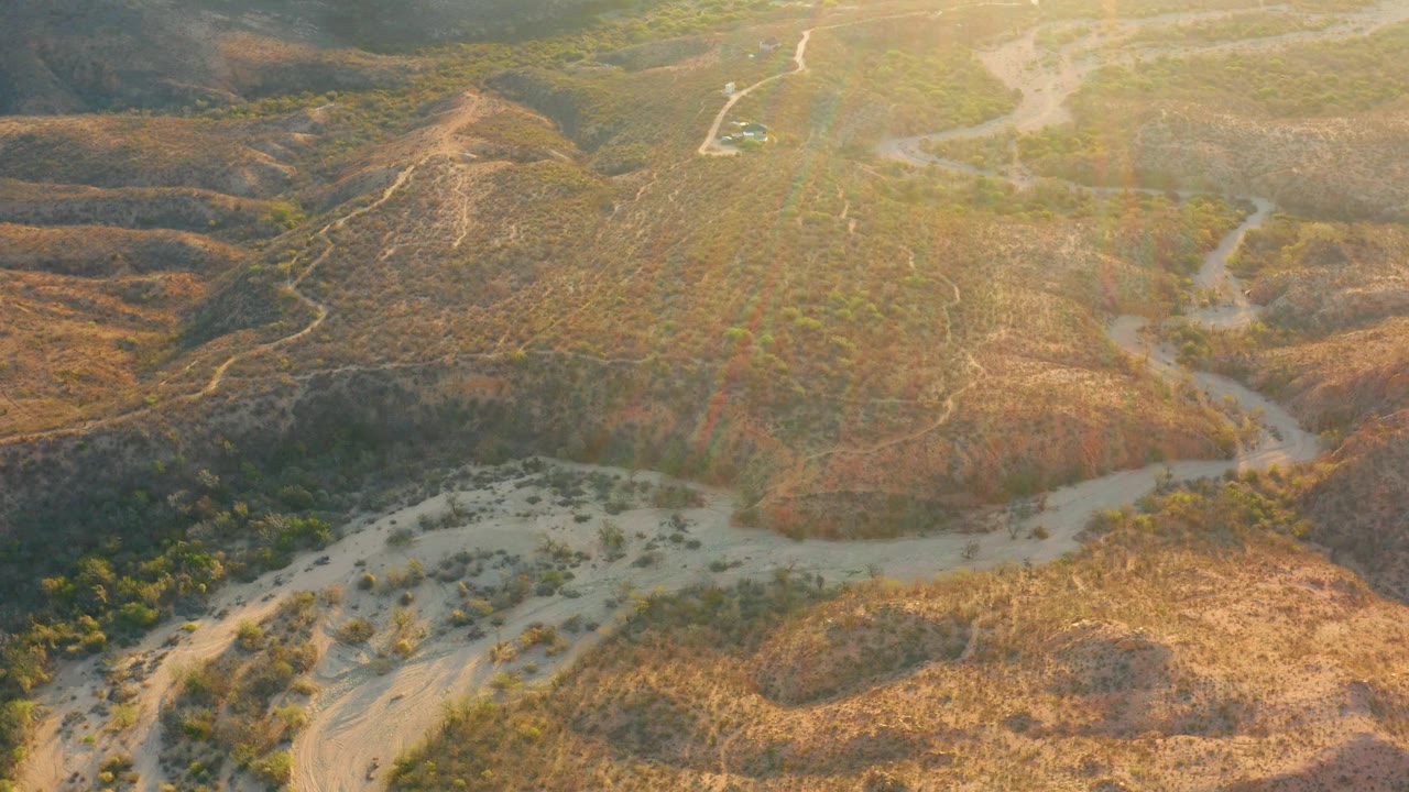 Aerial view in Cabo Pulmo with a winding sandy channel cutting through brown hills, darker shaded slopes, green shrub patches, and no visible people.