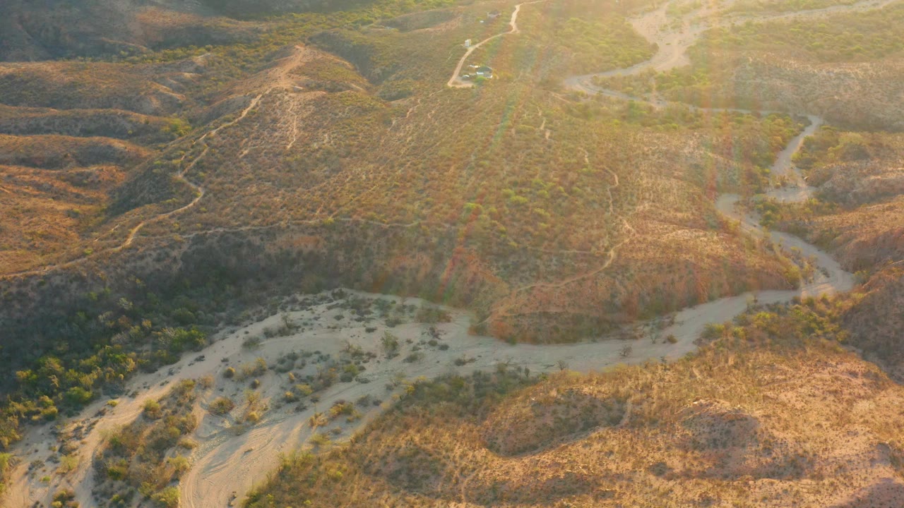 Drone-style aerial frame over Cabo Pulmo dry hills with a branching wash in the midground, scattered green vegetation, faint tracks, and no visible people.