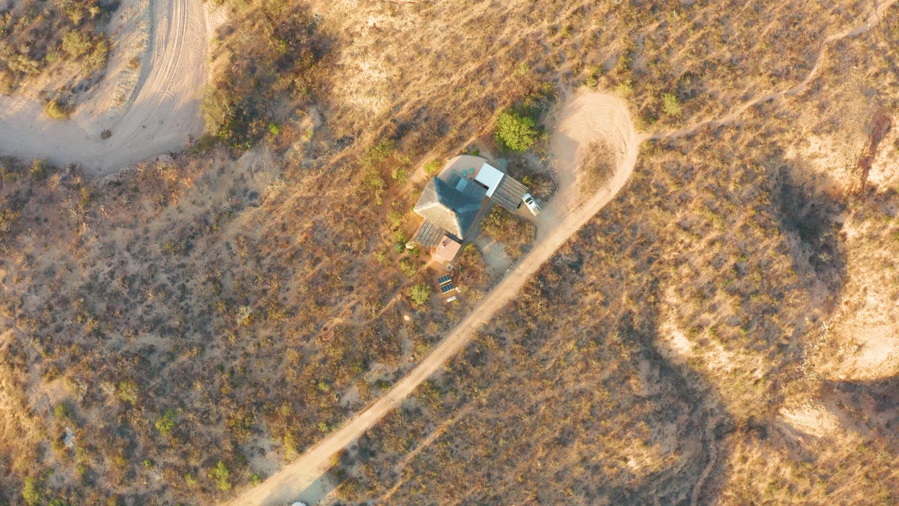 Aerial view over Cabo Pulmo dry scrubland with a pale dirt road curving past a small roofed building near the upper left, no visible people.