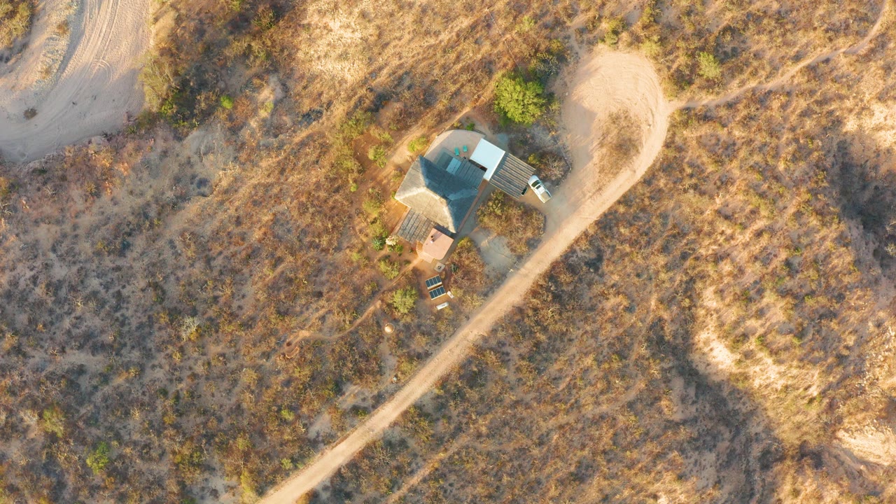 Early overhead frame of a rural house and curving dirt road in Cabo Pulmo, Baja California Sur, with dry grasses, scattered trees, and no visible people.