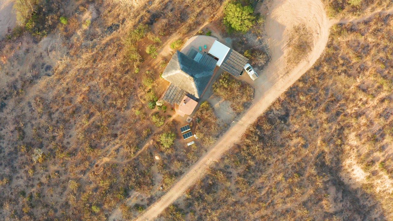 Overhead frame in Cabo Pulmo, Baja California Sur showing a small house, pale dirt road, dry grass, scattered shrubs, a light vehicle, and a tiny person near the structure.