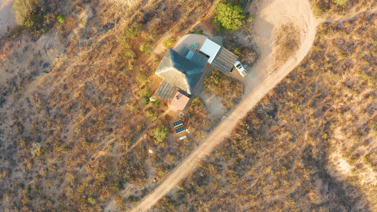 Overhead Cabo Pulmo frame with the roofed house near the upper left, a curving pale dirt road to the right, dry brown field, shrubs, and a tiny person near the house area.
