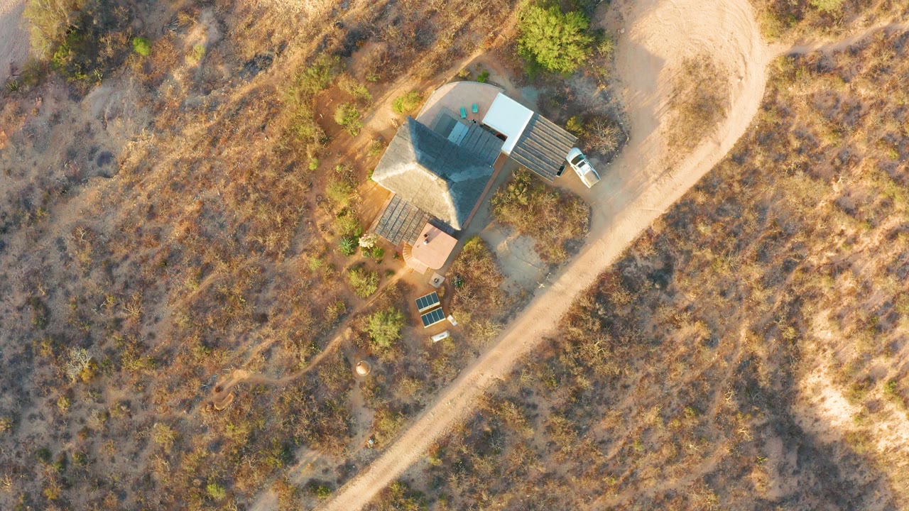 High overhead frame in Cabo Pulmo with a small rural house, parked light vehicle, pale dirt road, dry brushland, green vegetation patches, and a small person visible near the property.