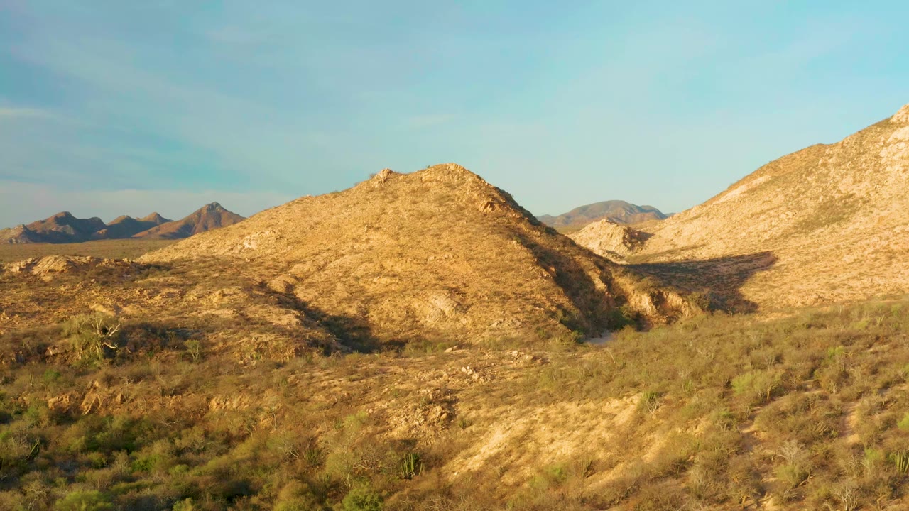Opening frame of Cabo Pulmo desert hills in Baja California Sur, Mexico, with low scrub in the foreground, a rounded tan hill at center, distant ridges at left, and no visible people.