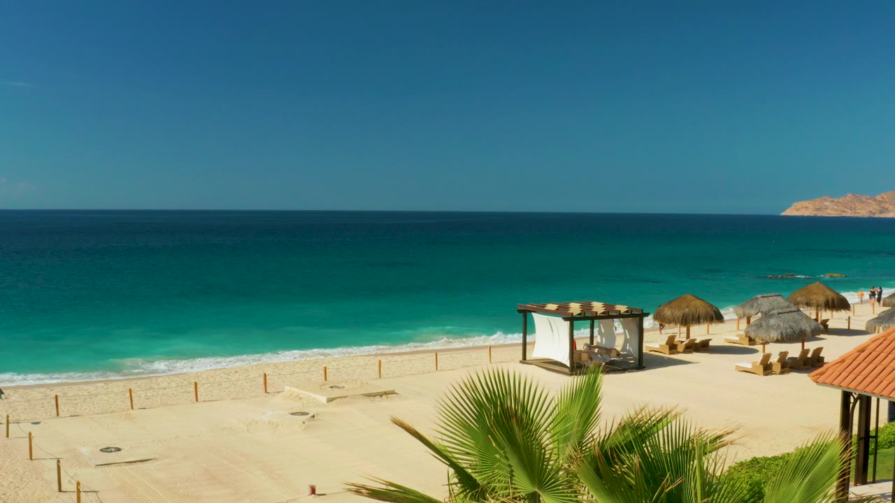 Hilton Los Cabos beach view with turquoise water, pale sand, thatched umbrellas, a white canopy structure, foreground palm fronds, and no visible people at the start of the clip.