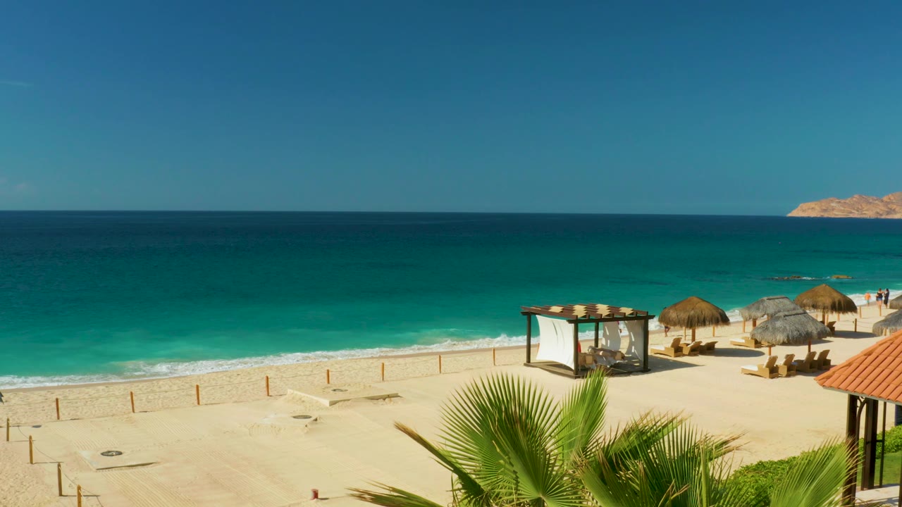 Hilton Los Cabos shoreline with bright turquoise surf, open sand, thatched umbrellas, a canopy structure, and palm fronds lower in the frame, with no visible people.